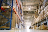 Imagem mostra corredor de galpão logístico com mercadorias dispostas em estantes industriais.