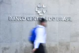 Imagem mostra rapaz de mochila caminhando em frente a parede com a logo e o nome do Banco Central do Brasil.