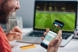 Rapaz feliz vibra com resultado de jogo de futebol na tela de aplicativo de aposta esportiva on-line em seu celular.