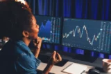 Jovem investidora negra comemora em frente a telas de computador com gráficos da Bolsa de Valores.