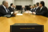 Placa em primeiro plano com a inscrição Copom e, desfocados no fundo, executivos engravatados sentados ao redor da mesa de reunião.