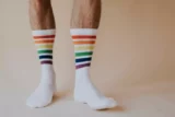 Imagem mostra detalhe de pés com meias brancas adornada por faixas coloridas em referência ao movimento LGBTQIA+