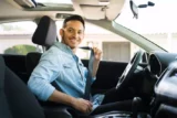 Imagem mostra jovem motorista homem colocando o cinto de segurança dentro do carro e exibindo um largo sorriso.