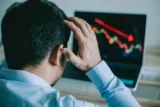 Homem de negócios visto de costas leva as mãos à cabeça em sinal de estresse enquanto olha para a tela do notebook, que mostra o gráfico do mercado financeiro caindo. Conceito de mercado de ações.