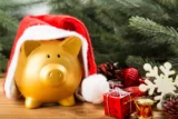 imagem de um cofre no formato de porquinho dourado com touca do papai noel ao lado de uma árvore de natal e presentes