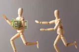Imagem mostra animação de bonequinhos de madeira roubando dinheiro: enquanto um foge com notas de ólares embaixo do braço, o outro corre atrás.