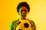 Imagem de fã de futebol afro-americana com bandeira do Brasil torcendo em iluminação amarela. (Foto: vectorfusionart em Adobe Stock)