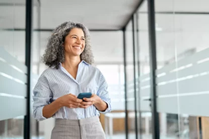 Mulher de negócios de meia idade feliz e sorridente segurando celular no corredor de um escritório.