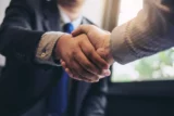 Imagem mostra detalhe de mãos de dois homens de negócios se apertando após fechamento de acordo, tornando-se parceiros.