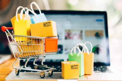 Imagem mostra carrinho de compras cheio de sacolas, na frente de uma tela de computador, fazendo alusão às compras online