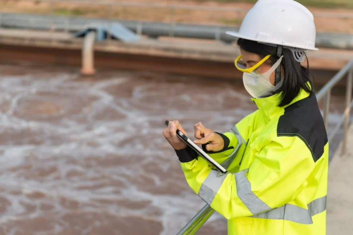 Dividendos de empresas de saneamento. Imagem mostra engenheira ambiental fazendo anotações em tablet durante trabalho em reservatório de estação de tratamento de água.