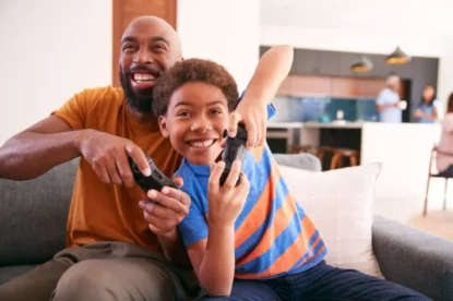 Pai e filho negros se divertem no sofá em casa jogando videogame juntos.