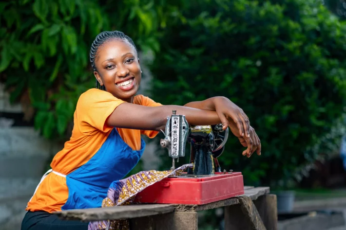 Imagem de uma mulher empreendedora africana sentada com as mãos em uma máquina de costura.