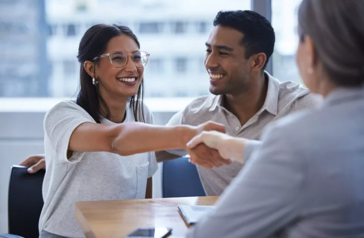 Imagem de um jovem casal apertando a mão de gerente do banco ao fechar acordo de financiamento imobiliário.