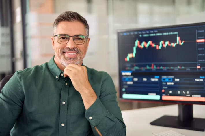 Investidor homem de meia idade sorri para retrato. Ele usa óculos, tem barba curta e grisalha e veste camisa. Ao fundo, monitor mostra gráficos da bolsa de valores.