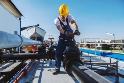 Trabalhador de macacão e capacete na cabeça maneja válvula de tubulação de petróleo sob céu azul.