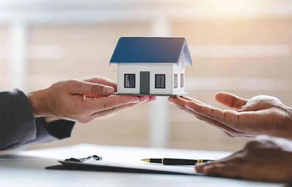 mãos de duas pessoas segurando uma casa em miniatura e um contrato com uma caneta sobre a mesa