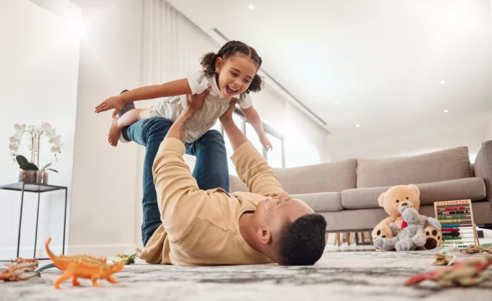 Família feliz, pai e menina brincando em uma casa com liberdade, criando laços e aproveitando tempo de qualidade juntos. Felicidade, sorriso e criança voando nos braços do pai deitado no chão. Conceito de independência financeira.