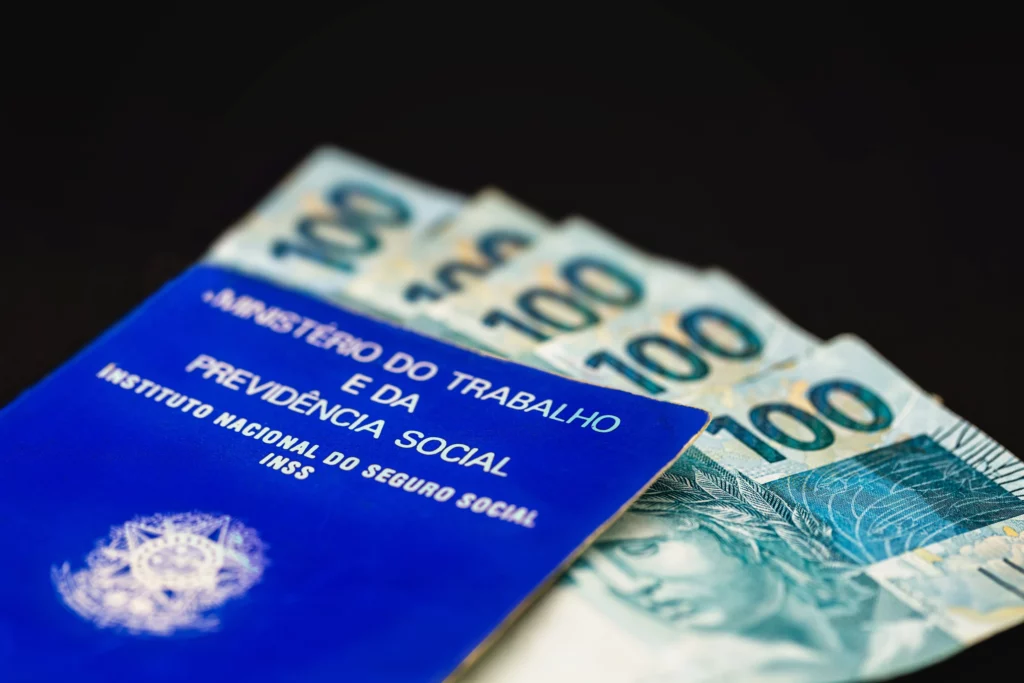 Foto mostra detalhe da capa da carteira de trabalho brasileira sobre notas de 100 reais.