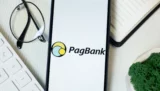 Tela de smartphone com uma imagem da logo do PagBank. O PagBank encerrou o primeiro trimestre de 2026 com lucro líquido de R$ 575 milhões e 34 milhões de clientes. (Imagem: Adobe Stock)