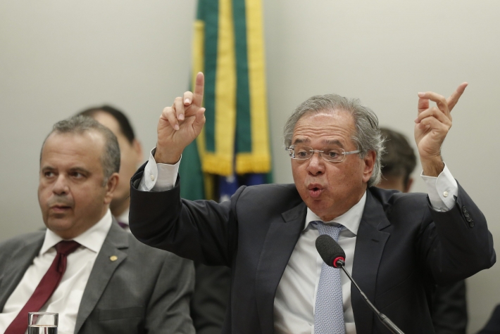 Paulo Guedes ergue as duas mãos, com os indicadores para o alto, enquanto fala durante sessão na Câmara. À sua direita está Rogério Marinho.