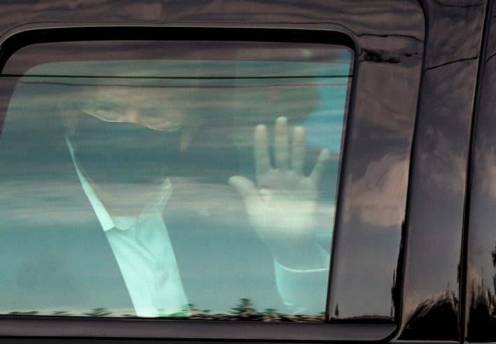 Donald Trump, vestindo máscara, acena de dentro de um carro com vidro fechado.